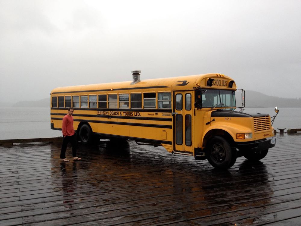 The yellow school bus parked on a dock by the water in the rain, with the chimney visible on the roof